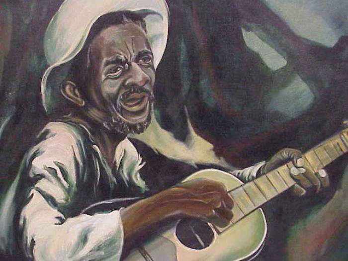 An older Black man in a white shirt and white hat hold a guitar and sings.